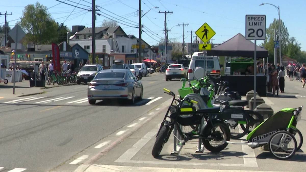 Street scene with bright green bicycles parked at a curb, market stalls and pedestrians along a sunny downtown road as cars drive by.