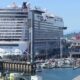 Huge white cruise ship Norwegian Encore docked at a busy harbor, with boats and a marina in the foreground and calm blue water.