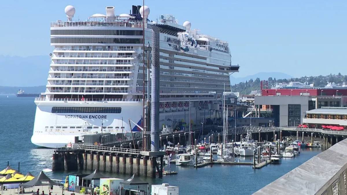 Huge white cruise ship Norwegian Encore docked at a busy harbor, with boats and a marina in the foreground and calm blue water.