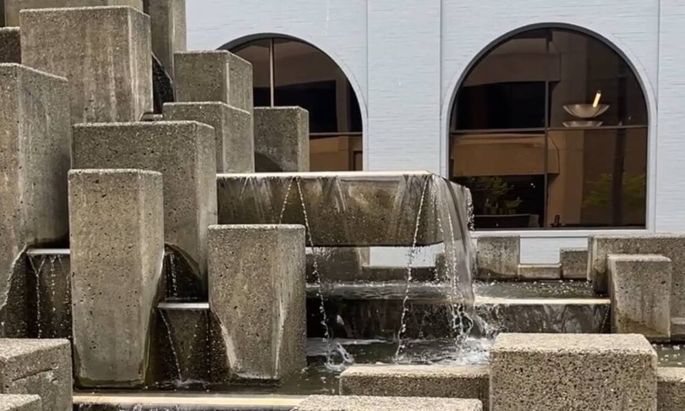 Tiered concrete fountain with water cascading over blocks in front of a blue building with arched windows.