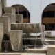 Tiered concrete fountain with water cascading over blocks in front of a blue building with arched windows.