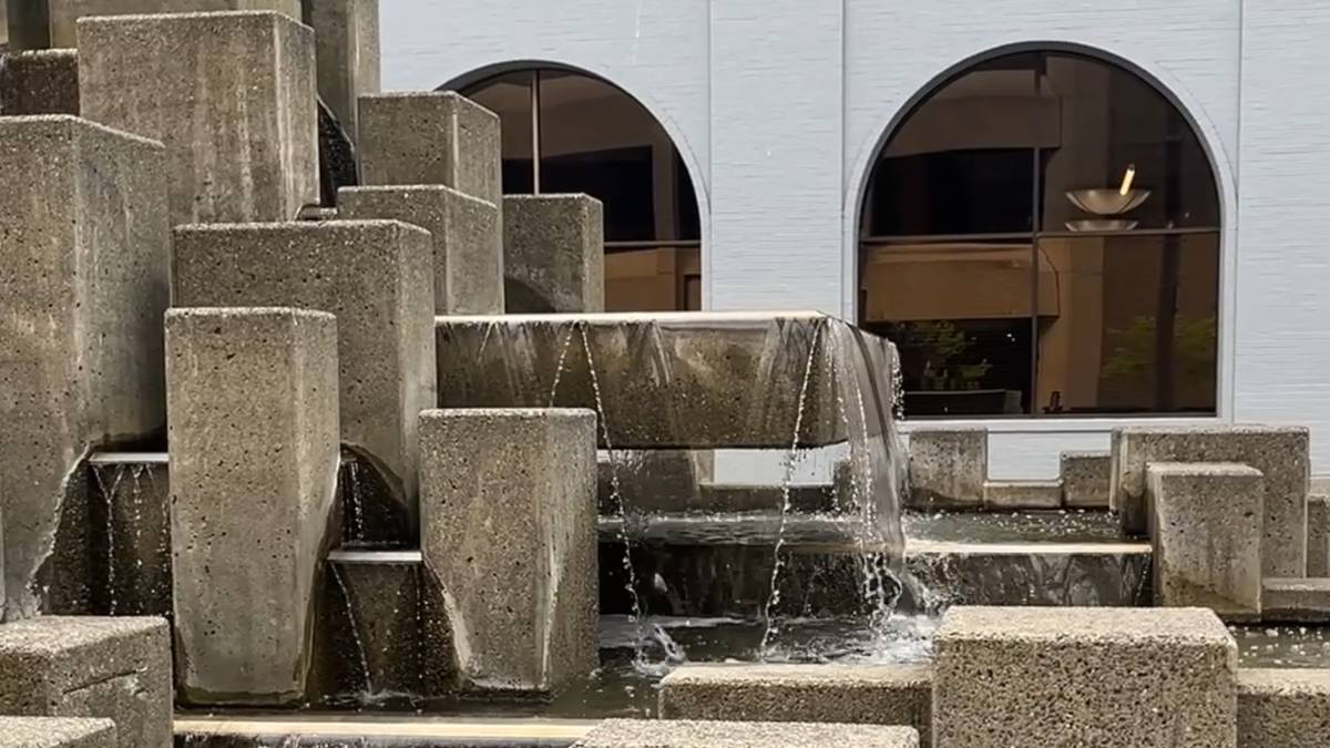 Tiered concrete fountain with water cascading over blocks in front of a blue building with arched windows.