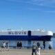Large blue-and-white cargo ship named GLOVIS on calm water, with people walking along a waterfront promenade in the foreground.