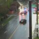 Rain-soaked city street with a black sedan in the center lane and a police car with flashing lights behind, reflections on the wet pavement, storefronts on the right including Chiang's Gourmet.