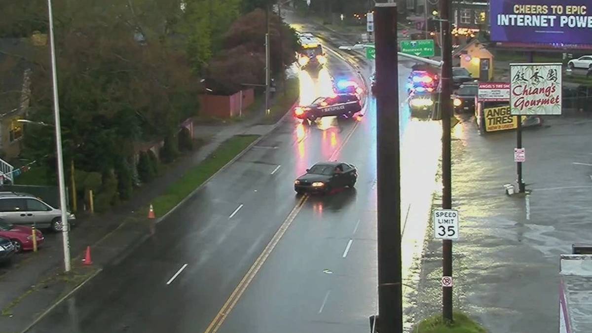 Rain-soaked city street with a black sedan in the center lane and a police car with flashing lights behind, reflections on the wet pavement, storefronts on the right including Chiang's Gourmet.