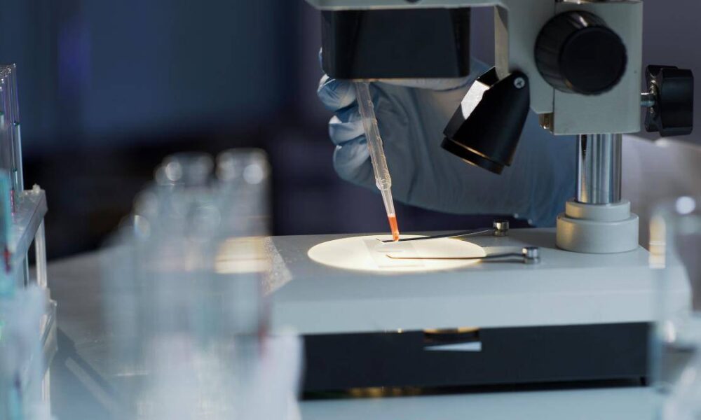 Gloved hand with a pipette placing a drop of liquid on a microscope slide under a microscope in a laboratory setting.