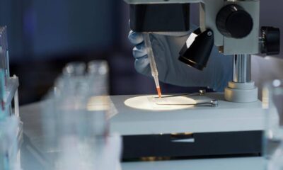 Gloved hand with a pipette placing a drop of liquid on a microscope slide under a microscope in a laboratory setting.