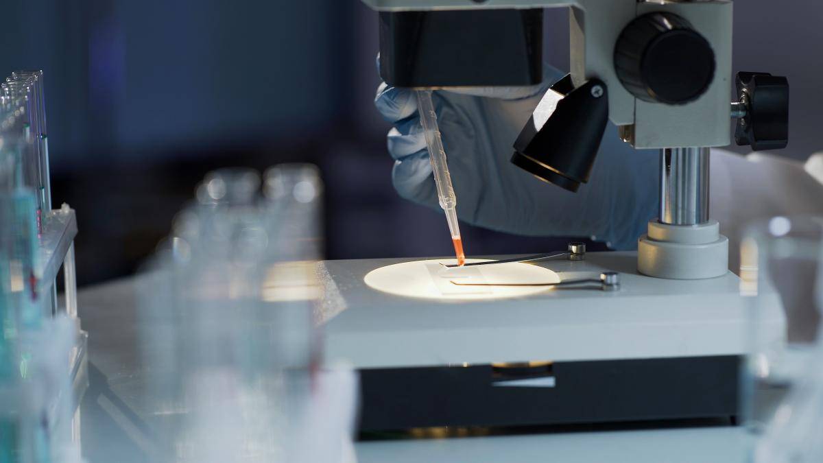 Gloved hand with a pipette placing a drop of liquid on a microscope slide under a microscope in a laboratory setting.
