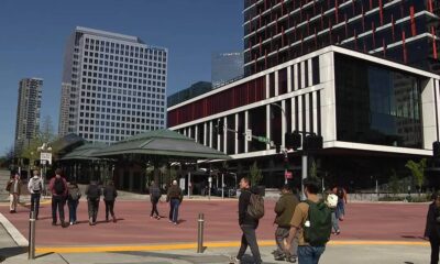 Group of people walking across a red-paved plaza beside a modern glass and white-framed building under construction in a city center.