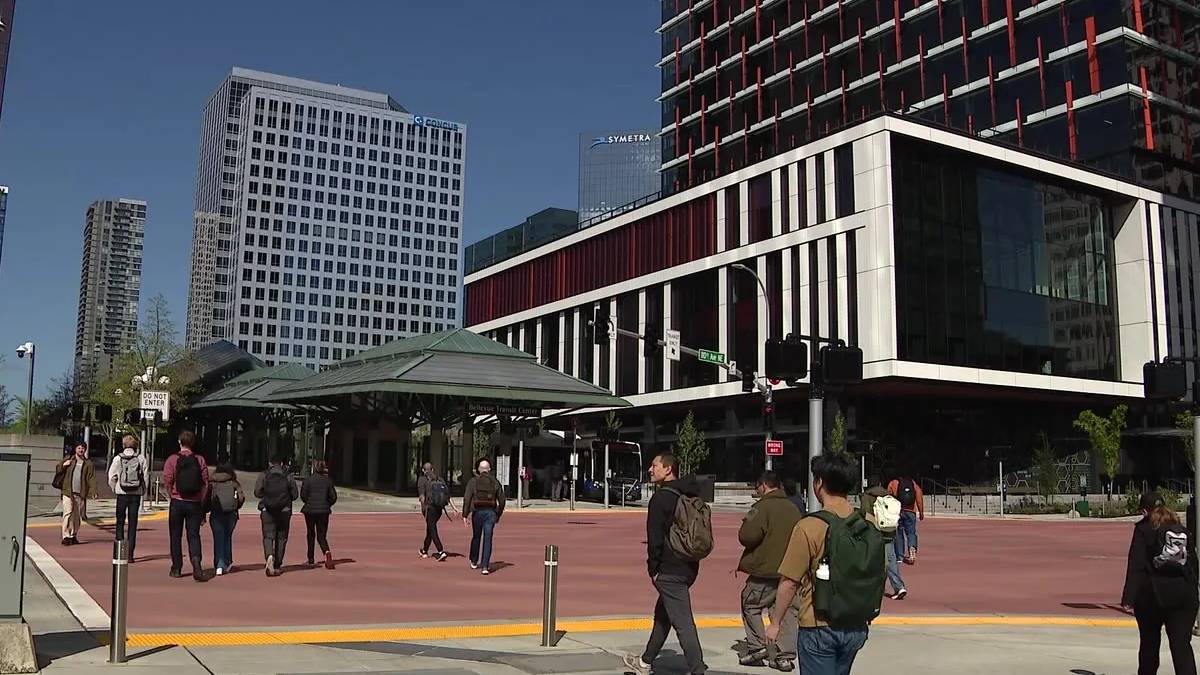 Group of people walking across a red-paved plaza beside a modern glass and white-framed building under construction in a city center.