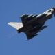 Fighter jet photographed from below as it climbs against a clear blue sky, viewed from beneath.