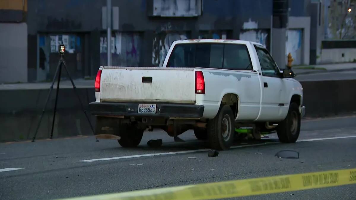White pickup truck on a city street behind yellow caution tape, with a tripod-mounted camera set up nearby behind it.