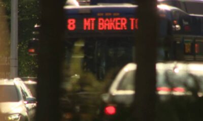 Public bus on a city street, red LED sign reads '8 MT BAKER...' visible through tree trunks in the foreground.