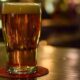 A pint of amber beer with a foamy head on a wooden bar, resting on a red coaster.
