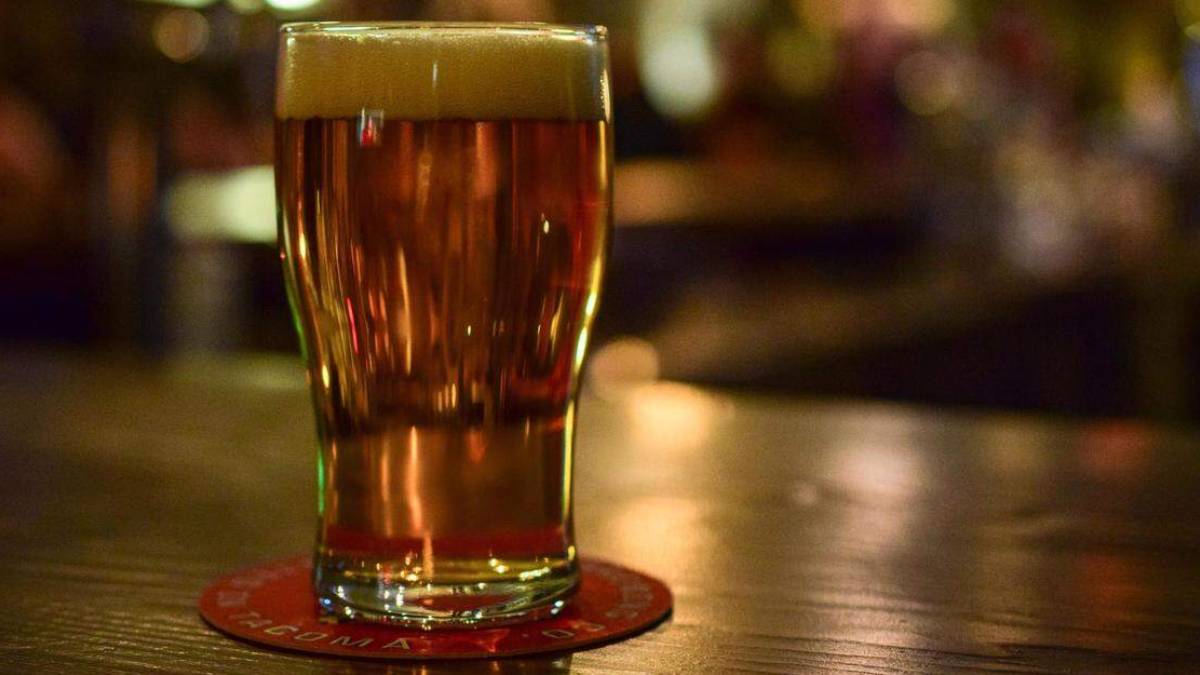 A pint of amber beer with a foamy head on a wooden bar, resting on a red coaster.