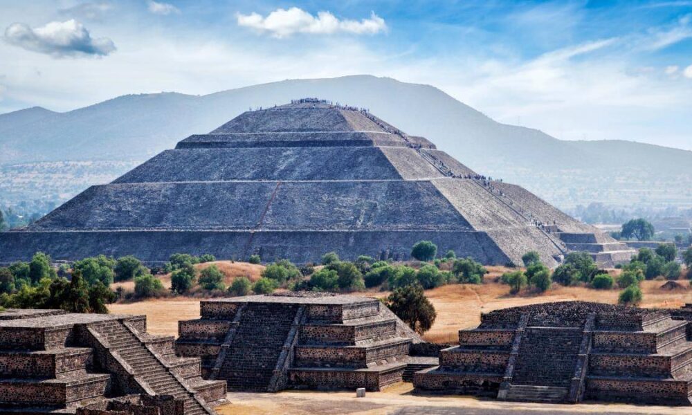 Massive stepped pyramid at Teotihuacan with a long central stairway and surrounding smaller pyramids set against hills and a blue sky.