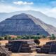 Massive stepped pyramid at Teotihuacan with a long central stairway and surrounding smaller pyramids set against hills and a blue sky.