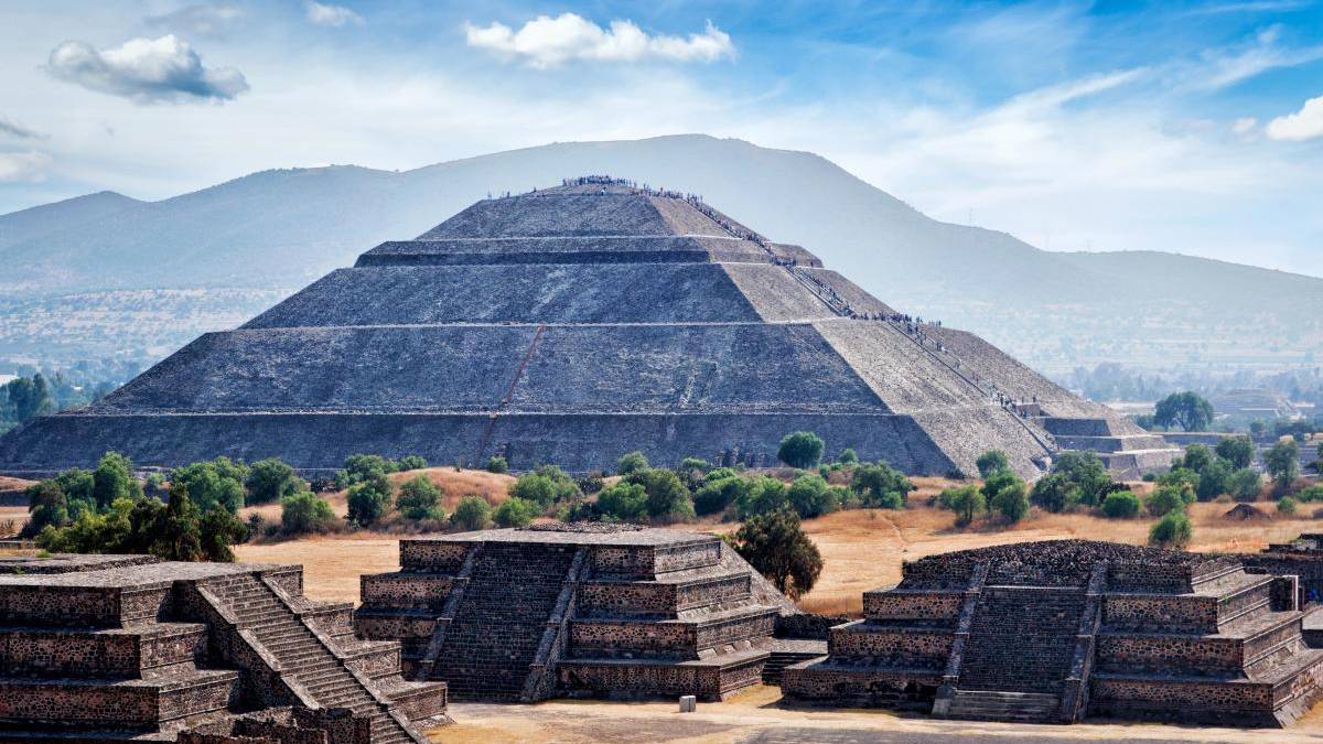 Massive stepped pyramid at Teotihuacan with a long central stairway and surrounding smaller pyramids set against hills and a blue sky.