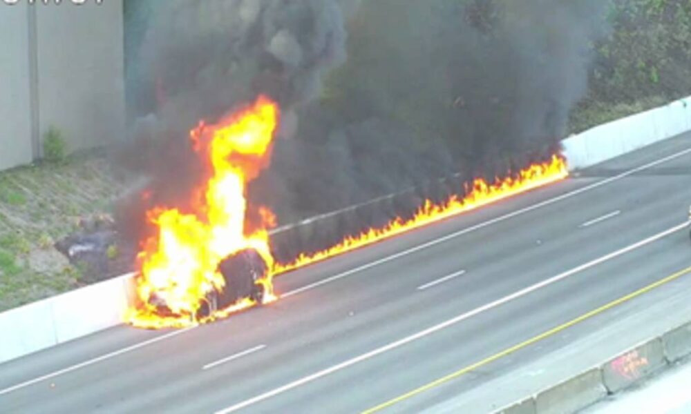 Car on a highway engulfed in bright flames with black smoke rising, flames along the guardrail as traffic passes nearby (no responders visible).