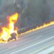 Car on a highway engulfed in bright flames with black smoke rising, flames along the guardrail as traffic passes nearby (no responders visible).