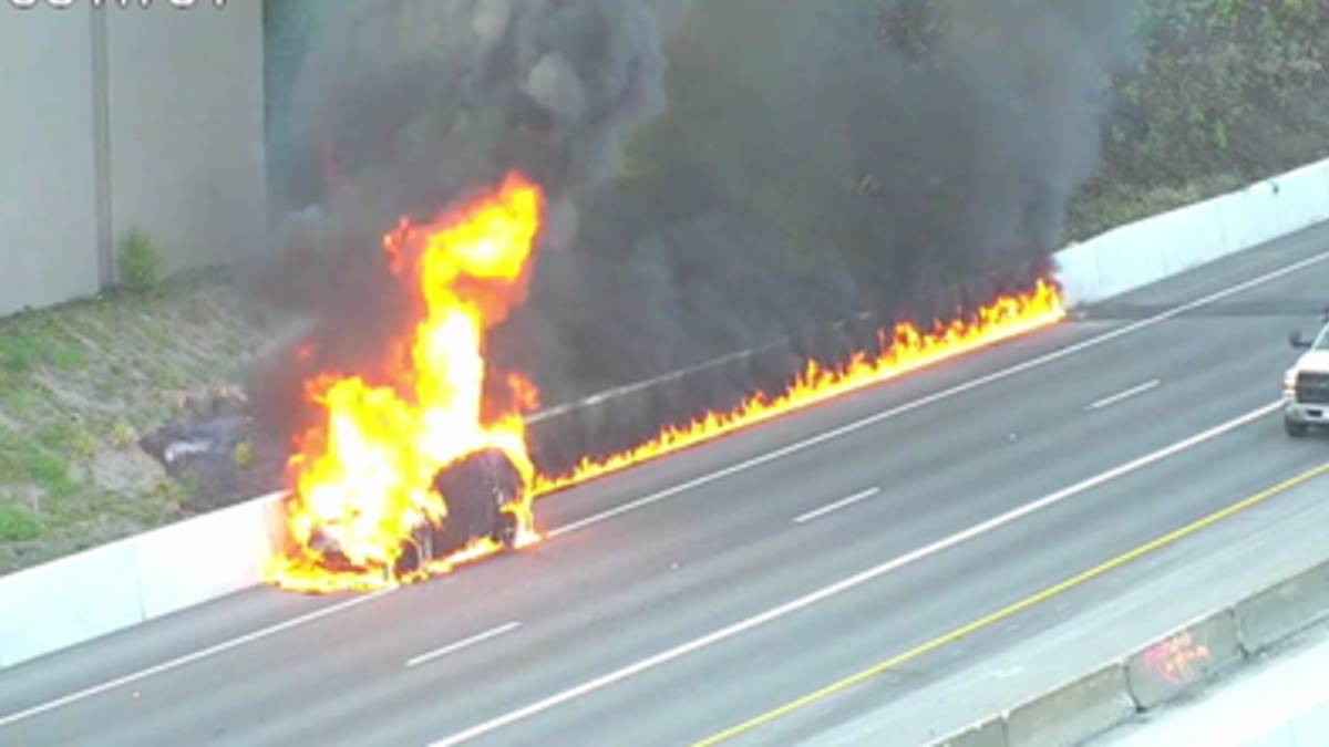 Car on a highway engulfed in bright flames with black smoke rising, flames along the guardrail as traffic passes nearby (no responders visible).