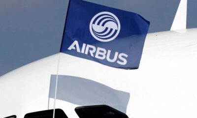 Blue Airbus flag with white logo fluttering near the tail of a white commercial airplane, casting a shadow on the fuselage.