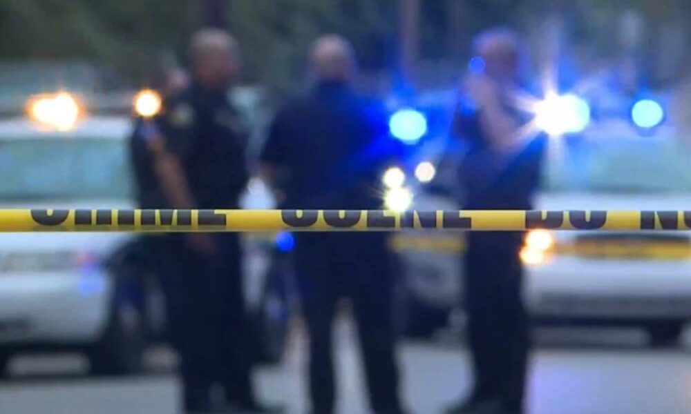 Yellow police tape in foreground with blurred officers and police cars with flashing lights in the background, indicating a crime scene or investigation area.