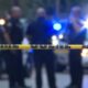 Yellow police tape in foreground with blurred officers and police cars with flashing lights in the background, indicating a crime scene or investigation area.