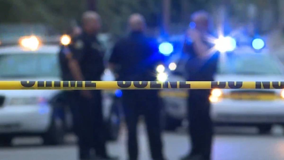 Yellow police tape in foreground with blurred officers and police cars with flashing lights in the background, indicating a crime scene or investigation area.