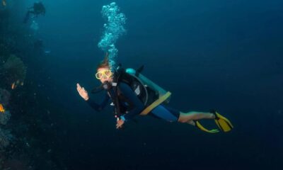 Scuba diver with yellow mask swimming underwater near a coral reef and bubbles.Ordinary scuba gear in blue water near coral growth, waving to the camera.