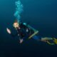Scuba diver with yellow mask swimming underwater near a coral reef and bubbles.Ordinary scuba gear in blue water near coral growth, waving to the camera.