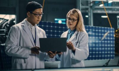Two scientists in white lab coats review data on a laptop in a solar panel research facility above them in the background.