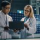 Two scientists in white lab coats review data on a laptop in a solar panel research facility above them in the background.