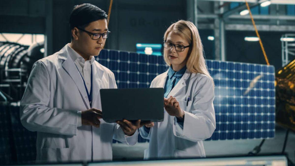 Two scientists in white lab coats review data on a laptop in a solar panel research facility above them in the background.
