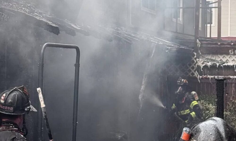 Firefighters in turnout gear spraying water in a smoky backyard fire scene.