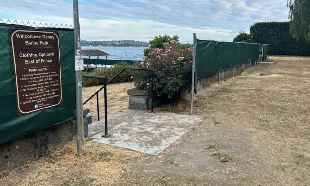 Entrance to Denny Blaine Park by the lake with a brown informational sign reading 'Clothing Optional: East of Fence' near a railing and concrete pad.