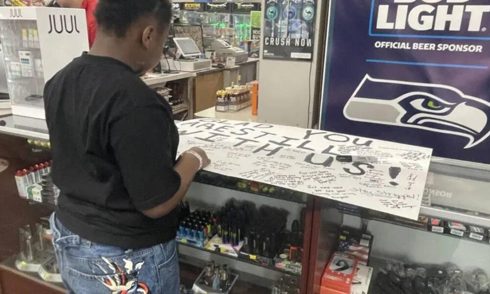 A man in a black shirt signs a large white board covered in signatures at a store counter, with a Seattle Seahawks banner in the background.