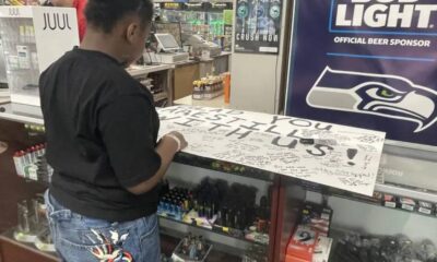 A man in a black shirt signs a large white board covered in signatures at a store counter, with a Seattle Seahawks banner in the background.