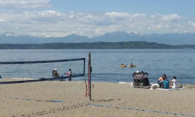 Beach volleyball net on a sandy shore with people sitting nearby, calm water and distant mountains under a blue sky.