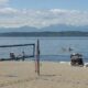 Beach volleyball net on a sandy shore with people sitting nearby, calm water and distant mountains under a blue sky.