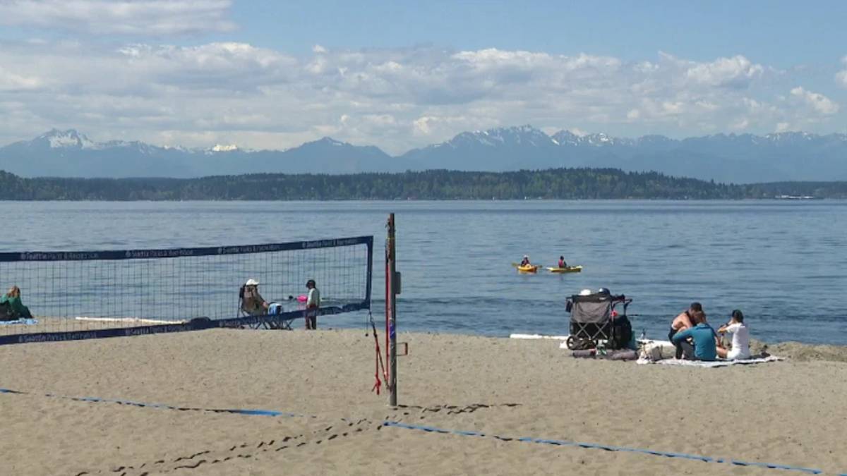 Beach volleyball net on a sandy shore with people sitting nearby, calm water and distant mountains under a blue sky.