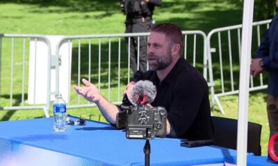 Bearded man speaks into a handheld microphone at a blue outdoor table, with a camera on a tripod facing him.
