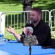 Bearded man speaks into a handheld microphone at a blue outdoor table, with a camera on a tripod facing him.
