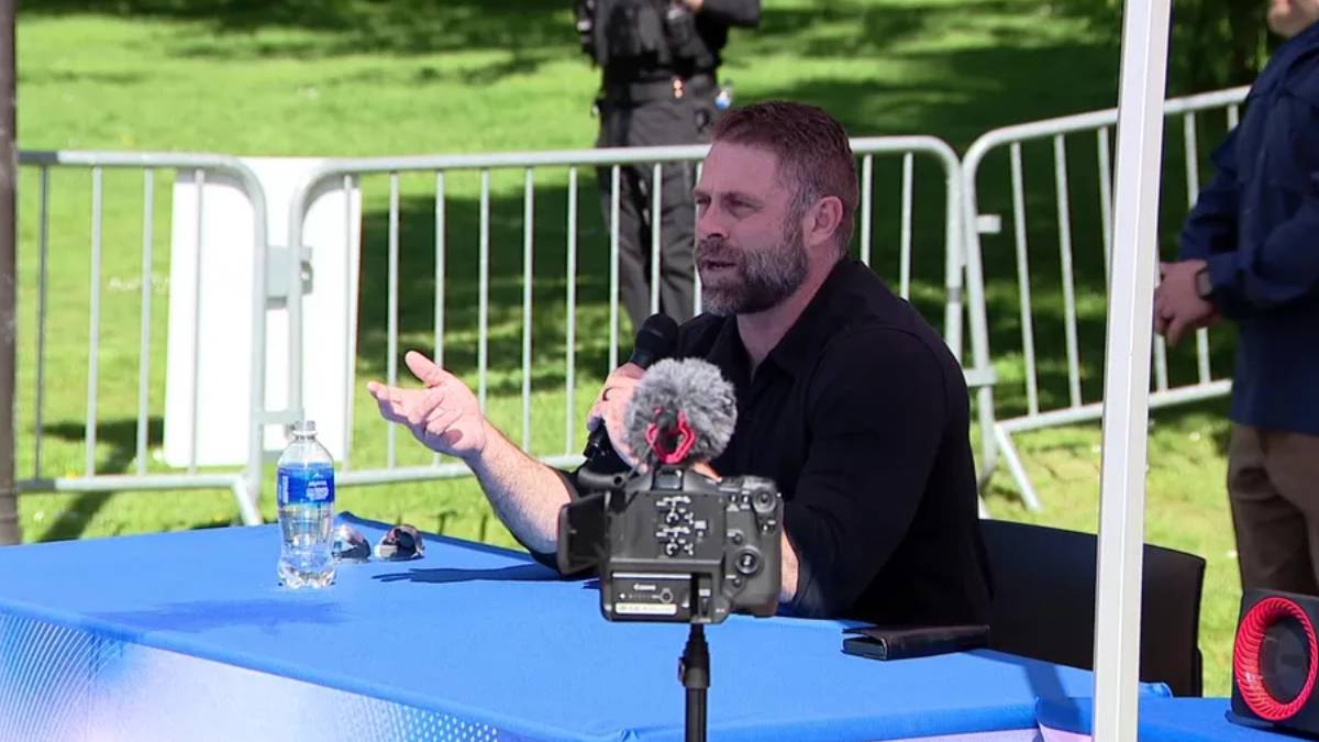 Bearded man speaks into a handheld microphone at a blue outdoor table, with a camera on a tripod facing him.