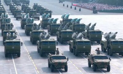 Ranked rows of green missile-carrying trucks in a military parade on a wide runway, with soldiers nearby.