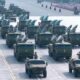 Ranked rows of green missile-carrying trucks in a military parade on a wide runway, with soldiers nearby.