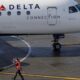 Delta Connection regional airliner on the tarmac, ground crew member in a bright vest signaling with a baton nearby.