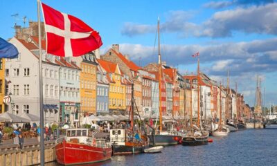 Colorful waterfront houses along a canal with moored boats in Nyhavn, Copenhagen; a Danish flag waves in the foreground.
