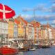 Colorful waterfront houses along a canal with moored boats in Nyhavn, Copenhagen; a Danish flag waves in the foreground.