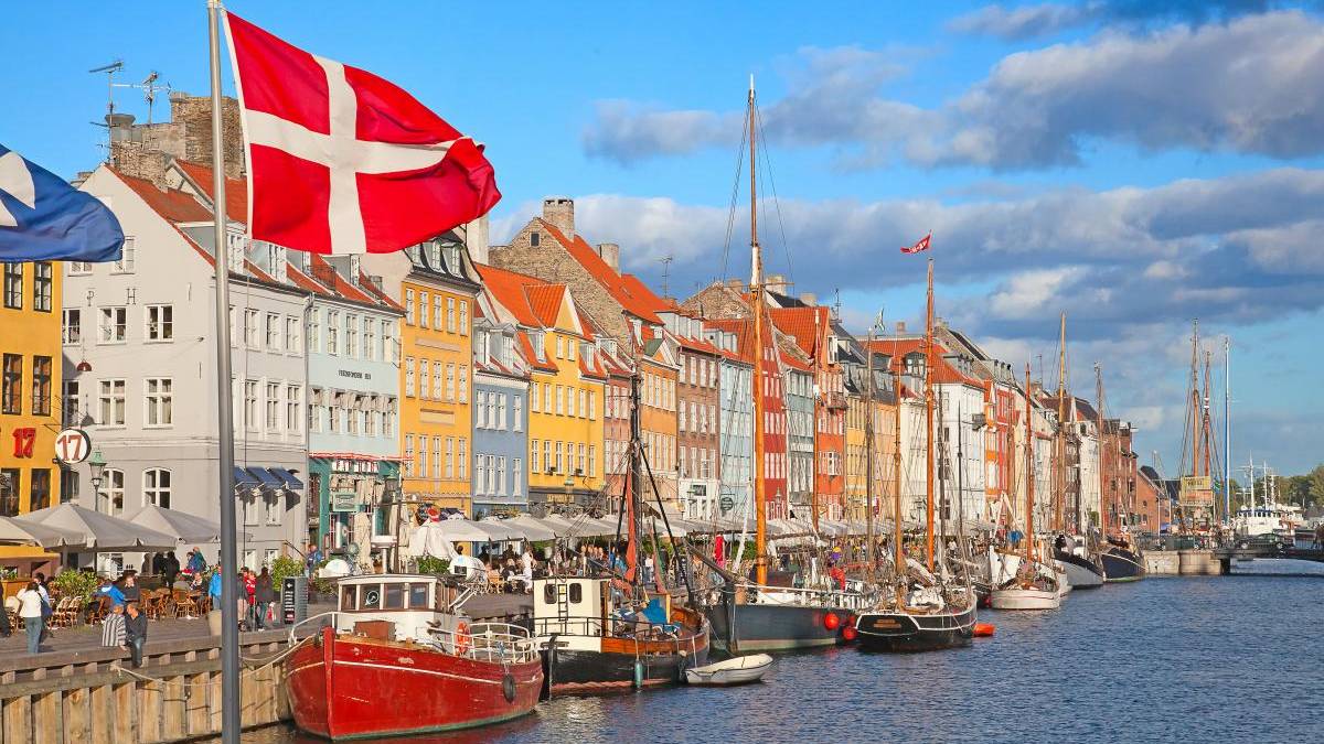 Colorful waterfront houses along a canal with moored boats in Nyhavn, Copenhagen; a Danish flag waves in the foreground.